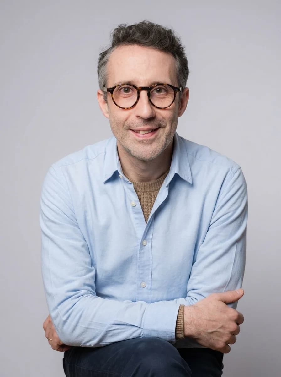 Portrait IA dentiste style corporate chaleureux - chemise bleu clair sur pull col rond beige, posture détendue bras croisés, fond neutre